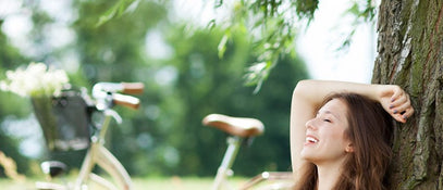 A woman sitting against a tree with her hand over her head and her bicycle in the background