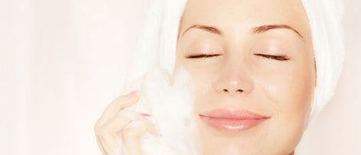 A woman smiling with her eyes closed, wearing a white turban and washing the side of her face with soap